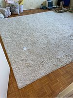 White area rug laid flat on wooden parquet floor, showing the overall size, rectangular shape, and texture of the rug. Some items are visible around the room edges.