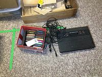 Atari 2600 console with multiple cables and a box of game cartridges on carpeted floor