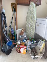 Gray and blue Eureka canister vacuum with hose attached, surrounded by assorted cleaning supplies including spray bottles, gloves, cleaning liquids, shoe mat, plastic bucket, and ironing board.