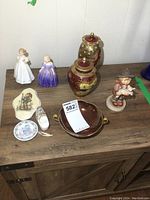 Overview of all items on wooden cabinet including two Royal Doulton figures, Carlton Ware urns and bowl, Hummel figurine, and other porcelain pieces.