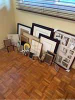 A group of over twenty assorted photo frames of various materials and sizes arranged on the floor by a window with wood blinds.