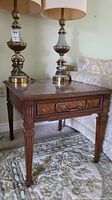 Front angle of the heritage wooden end table showing carved legs and drawer pull, with two lamps on top.