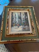 Framed painting showing a street-like courtyard scene with several dancers in colorful dresses between tall gray buildings. The frame is wide with gold and green hues.