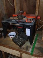 Overall view of router table on wooden bench with orange router and safety covers, includes open brown case of router bits in front.
