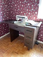 Kenmore sewing machine placed on top of a dark wood sewing cabinet with three drawers, side pull-down extension is open, floral wallpaper in background.