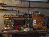 Overview of workbench with wooden chest, plastic organizers, hand tools, clamps, and power tools along back wall shelves