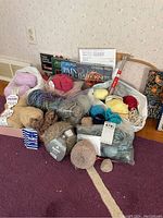 Wide view of five bags with yarn in various colors and thickness, knitting needles, wooden stretcher, and crafting instruction books.