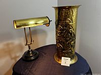 Brass bankers lamp and embossed brass umbrella stand side by side on dark cloth.