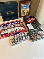 Photo shows 5 board games: Trivial Pursuit Master Game, Pari Parcheesi in wooden box, two Rummoli boxes, Uno Deluxe card game in a black box, and the Autobridge game. Also shows six new decks of playing cards at front.