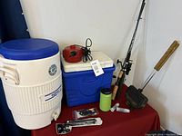 Wide view showing large Igloo water jug with spigot, blue Coleman cooler, red electric pump, two fishing rods, Thermacell repellent device, and panini press on table.