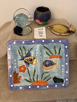 Lot items displayed on beige cloth: glass fish figurine, candle holder, yellow fish trinket dish, and hand-painted serving platter.