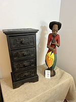 Wood carved cabinet with four drawers next to a wood carved fisherman statue holding a fish, both showing cracks