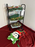 Photo showing black metal craft rack on wheels with three wire baskets filled with folded holiday fabric, and penguin-shaped ribbon holder on red surface.