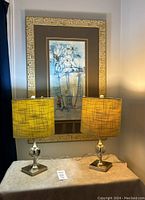 Photo showing large framed floral art piece with two table lamps having yellow fabric shades and metallic bases on a small table.