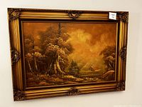 Front view of a large antique landscape painting housed in an ornate golden frame with detailed carvings at the corners. The scene shows tall trees and a cabin under a yellowish cloudy sky.