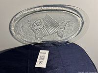 Top view of a clear glass fish-shaped serving platter showing detailed fish texture and bubbles. The platter is on a dark cloth background with a tag in front.