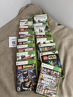 Xbox 360 game cases arranged on fabric surface showing titles such as LEGO Batman 2, LEGO Marvel, Assassin's Creed II, Grand Theft Auto V, and others.