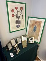 Two large green picture frames mounted on the wall each holding artwork, nine smaller frames arranged on a table beneath including gold-toned and checkered patterned frames