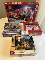 Four boxed 1000-piece puzzles showing front art including holiday train, childhood car dreams, Charlie Brown Christmas, and Big Ben village scene.
