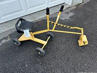 Side view of the yellow ride on sandbox digger crane showing the black seat, wheels, and two handles to operate the digging arm on asphalt ground in front of a white garage door.