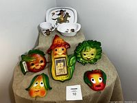 Front view of six colorful chalkware fruit plaques arranged on cloth, with Noritake kids dish set (tray, bowl, mug) on top right corner.