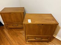 Pair of vintage wooden night tables, showing overall front and side views. Both have a top drop-down door and bottom drawer with wood handles, sitting on wooden legs, placed on hardwood floor.