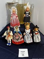 Front view of 6 collectible dolls in traditional European costumes on display cloth with tag showing lot number 15.