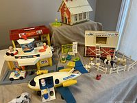 Wide view of all playsets including School House, Hospital, Family Farm, Airport playsets and various figures and accessories on gray cloth.