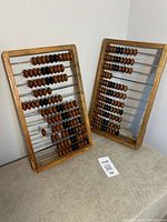 Two wooden abacuses leaning against a wall, showing wooden beads on metal rods within a wooden frame