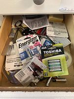Drawer containing various battery packages, loose batteries, metal can opener, bottle opener, packing material, and kitchen supplies.