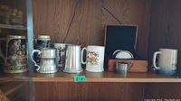 Full view of shelf with six beer steins and tankards including ceramic and metal varieties with some decorated scenes and plain finishes.