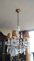 Full chandelier view showing six tan fabric shades, hanging clear crystals, brass metal frame, and glass column.