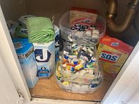Photo showing multiple plastic containers with dishwasher detergent pods, pack of green dish cloths and box of SOS steel wool pads inside a cabinet.