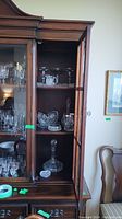 Tall wooden cabinet display containing various glass and crystal items including decanters and bowls.