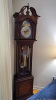 Full view of wooden grandfather clock showing ornate top, glass door with visible pendulum and weights, lower wood panel, and its overall tall stature.