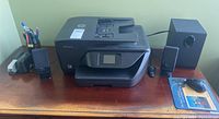Lot setup on wooden desk including HP OfficeJet 6962 printer, Edifier subwoofer and two speakers, mouse, stapler, and pen holder.