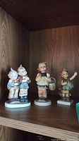 Three Hummel figurines displayed on wooden shelf.