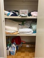 Linen closet shelf with assorted folded towels, bed sheets, quilts on top two shelves and cleaning supplies including spray bottles and a red dustpan on bottom shelf.