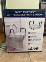Photo of the front of the box showing the raised toilet seat with removable padded arms, 5 inch height, and key features such as convenience, comfort, tool-free installation, and weight capacity 300 lbs.