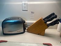 Photo of stainless steel toaster and wooden knife block with five knives on a kitchen counter.