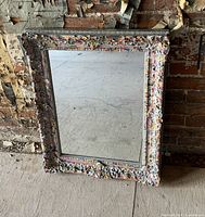 Photo of an antique carved wooden mirror with an ornate frame featuring splash paint in various colors, resting against a rustic brick wall.
