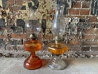 Two antique oil lamps set against a brick wall background. Both have glass bodies filled with orange oil and clear chimneys. The lamps vary slightly in design and base shape.