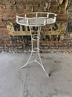 Full view of the antique Victorian wrought iron garden planter table painted white showing three-legged base, spool finials around circular tray top, set against a brick wall background.