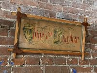 Front view of the framed punched paper needlework embroidered phrase 'Thine is the Kingdom', mounted on a brick wall. The wooden frame has decorative corner carvings and shows one leaf cluster missing.