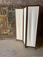 Photo of a 3-panel folding room divider with light wood frames and white linen panel inserts placed against an old brick wall.