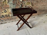 Wooden folding tray table with removable tray and foldable crossed-leg base placed on a concrete floor against a brick wall background.