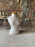 White female half-mannequin torso with wooden neck cap on a concrete floor against a brick wall.