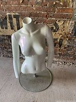 White female half torso mannequin with arms on round clear plexiglass stand against a textured brick wall background
