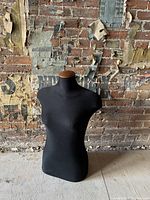 Black female mannequin torso against a partially worn brick and paper wall, on a floor with visible paint and minor dust.
