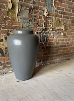 Large dark gray fibreglass garden urn standing on wood floor against a weathered brick wall.